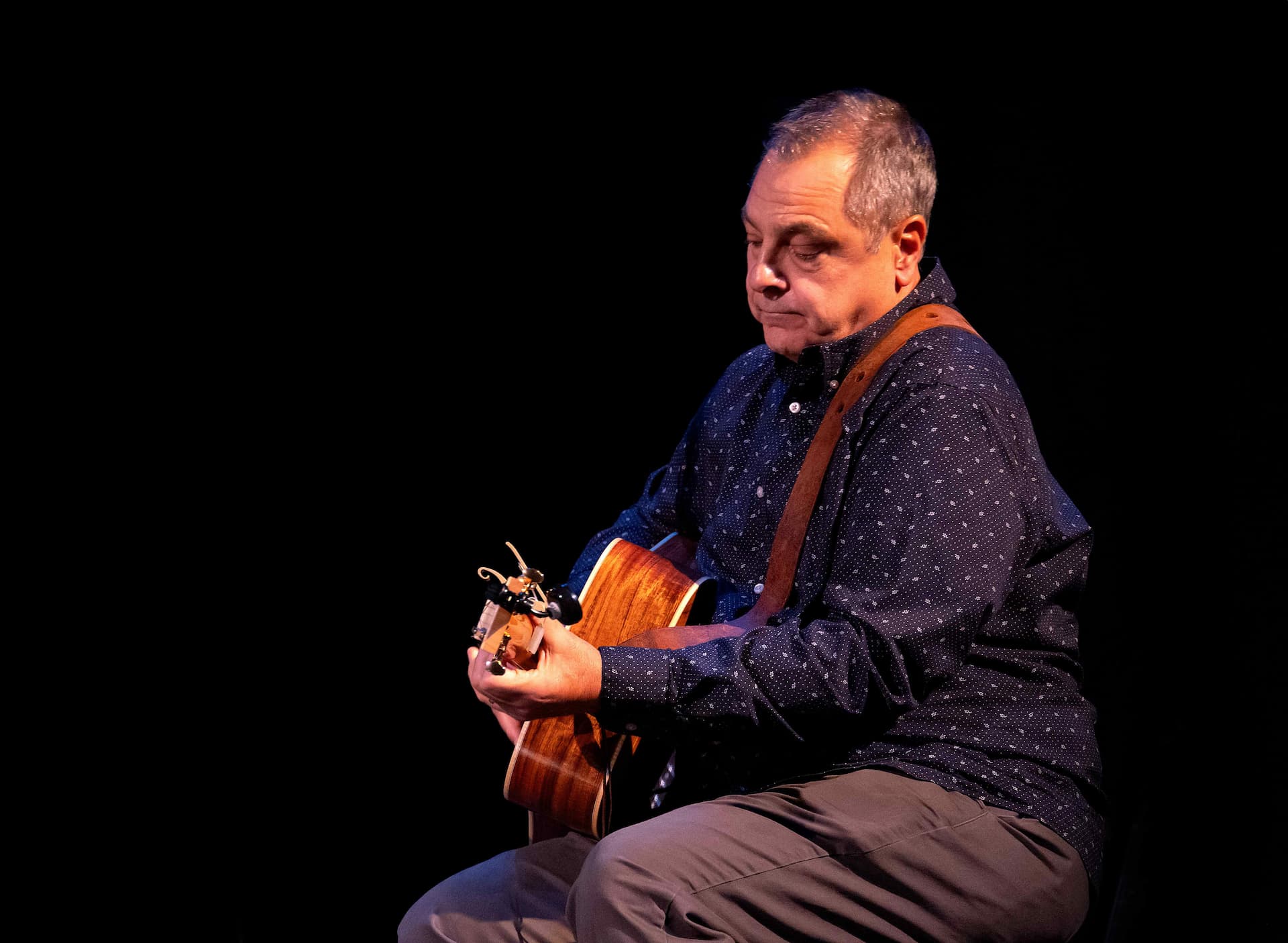 Your First Guitar Lesson  Singing  Guitar Lessons | Albany NY  Capital District | Peak Music A man sitting in the dark playing a guitar with the light focused on him